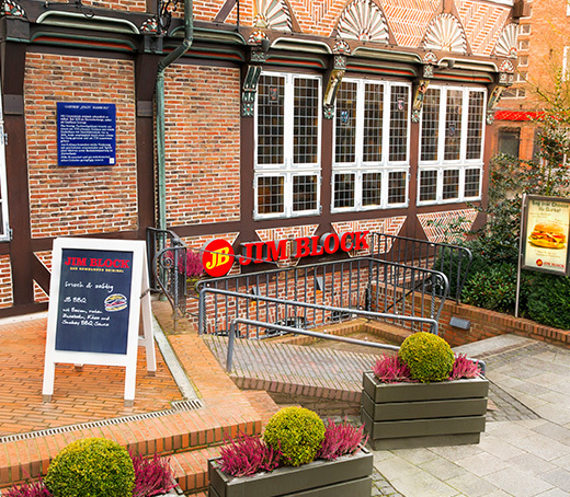 Eingang vom Jim Block Restaurant Bergedorf  Eingangsbereich des Jim Block Restaurants in Bergedorf mit Aufsteller, Pflanzen und Ziegelmauer.