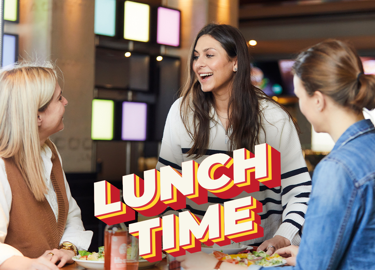 Jim Block Freunde essen Lunch Time  Drei junge Frauen essen gemeinsam im Jim Block Restaurant und lachen – Lunch Time Schriftzug im Vordergrund
