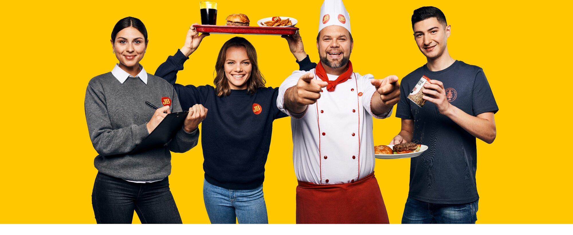 Jim Block Team Portrait Vier Jim Block Mitarbeitende in Uniform vor gelbem Hintergrund, zwei mit Speisen und Getränken, einer als Koch in Arbeitskleidung.