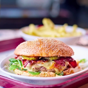 Ein saftiger Burger liegt auf einem Teller mit Sesambrötchen, gefüllt mit Käse, Speck, Gurken und frischem Gemüse. Im Hintergrund sind Pommes frites in einer Schüssel sichtbar.